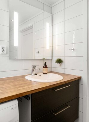 Ein Badezimmer mit einem weißen Waschbecken, einem Spiegel, einer Holzablage, einem dunklen Schrank und einer kleinen Pflanze.
