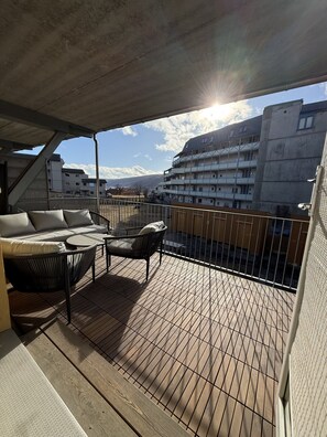 Un balcón con sofá y sillas, una terraza de madera y vistas a edificios al fondo.