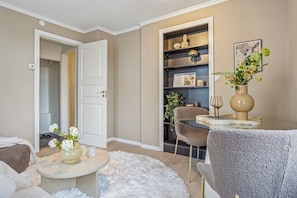 Una sala de estar con una puerta blanca, una mesa de vidrio, un sillón gris, un tapete blanco y una estantería con adornos.