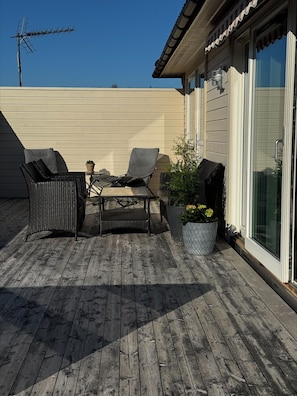 Une terrasse en bois avec deux chaises en osier et une table, une plante en pot et un bâtiment doté d’une fenêtre et d’une porte.