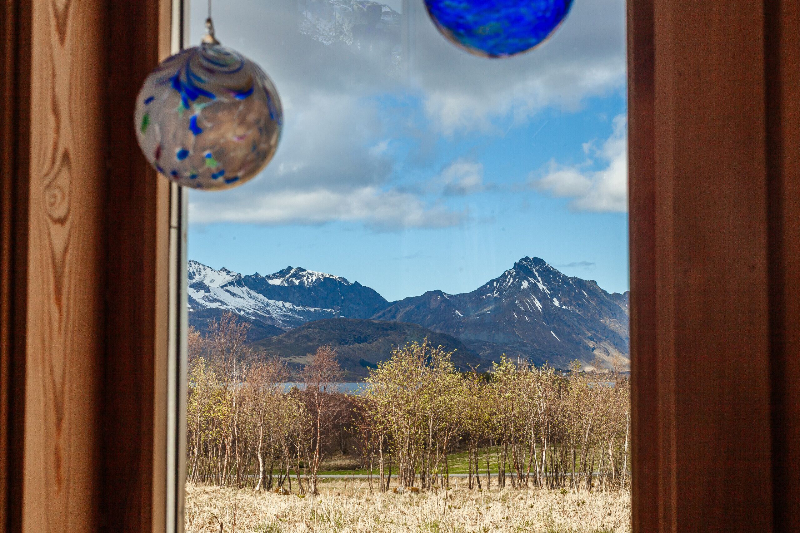 A view through a window with a wooden frame, featuring a mountainous landscape with snow-capped peaks and a clear sky.