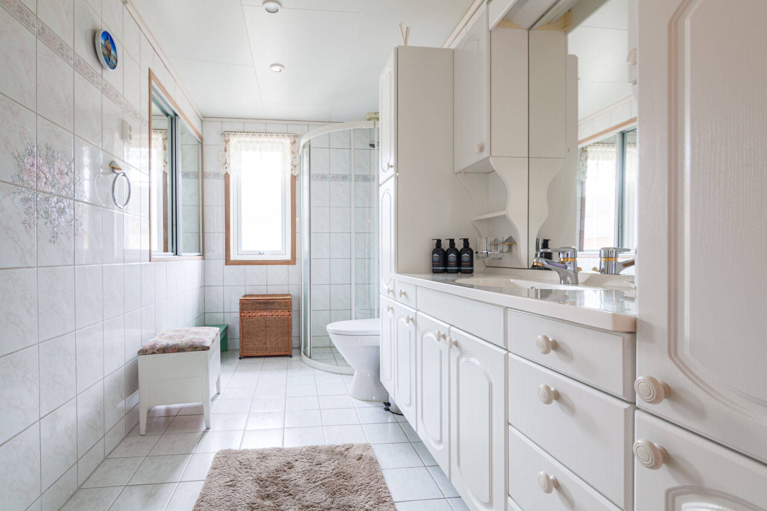 Un bagno rivestito di piastrelle bianche, con il water, una panca, un lavandino con delle bottiglie e uno specchio.