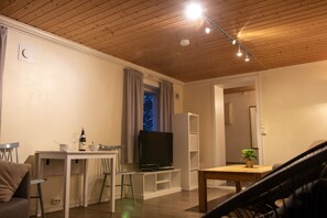 Una sala de estar con techo de madera, un televisor sobre un mueble bajo, una mesa de comedor con sillas y una planta en maceta.