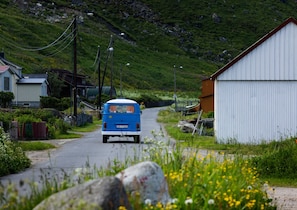 A blue van driving down a rural road with houses and a white building on the side.