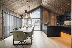 A modern kitchen with a wooden ceiling, a dining area with a long table and chairs, and a large window.