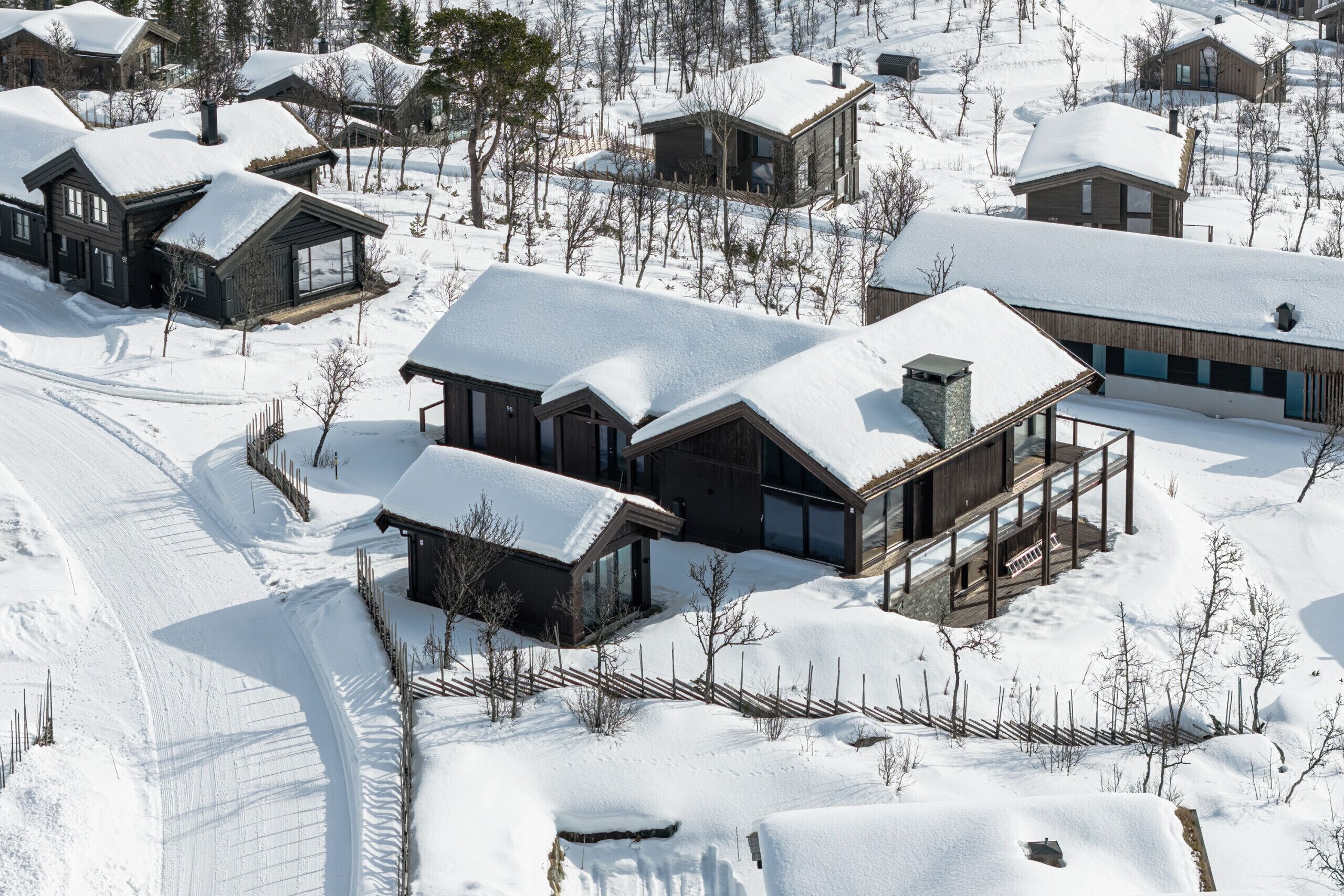 A snow-covered residential area with multiple houses, including a prominent wooden house with large windows and a balcony.