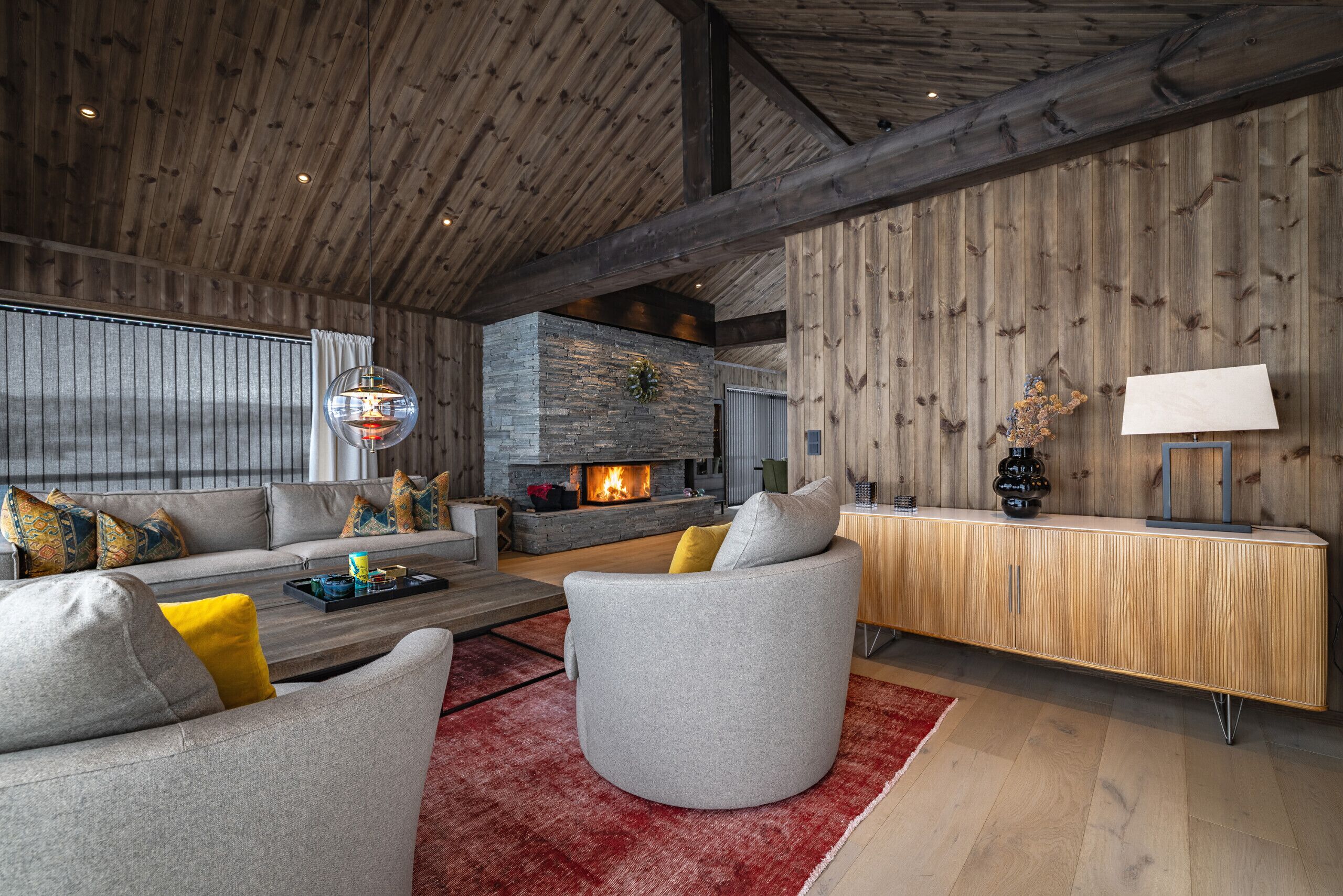 A modern living room with a stone fireplace, wooden ceiling beams, and a red rug.