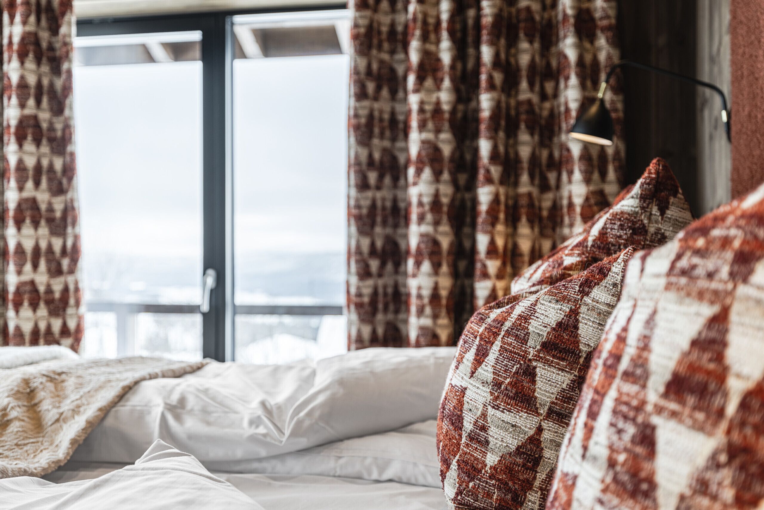 Una cama con sábanas blancas, una colcha roja y blanca a cuadros, y una ventana con cortinas.