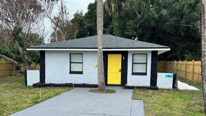 A small single-story house with a yellow door, a concrete driveway, and a wooden fence in the background. - Space Coast getaway! The Yellow Sunrise! 2 bed 1 bath Oasis (Merritt Island)