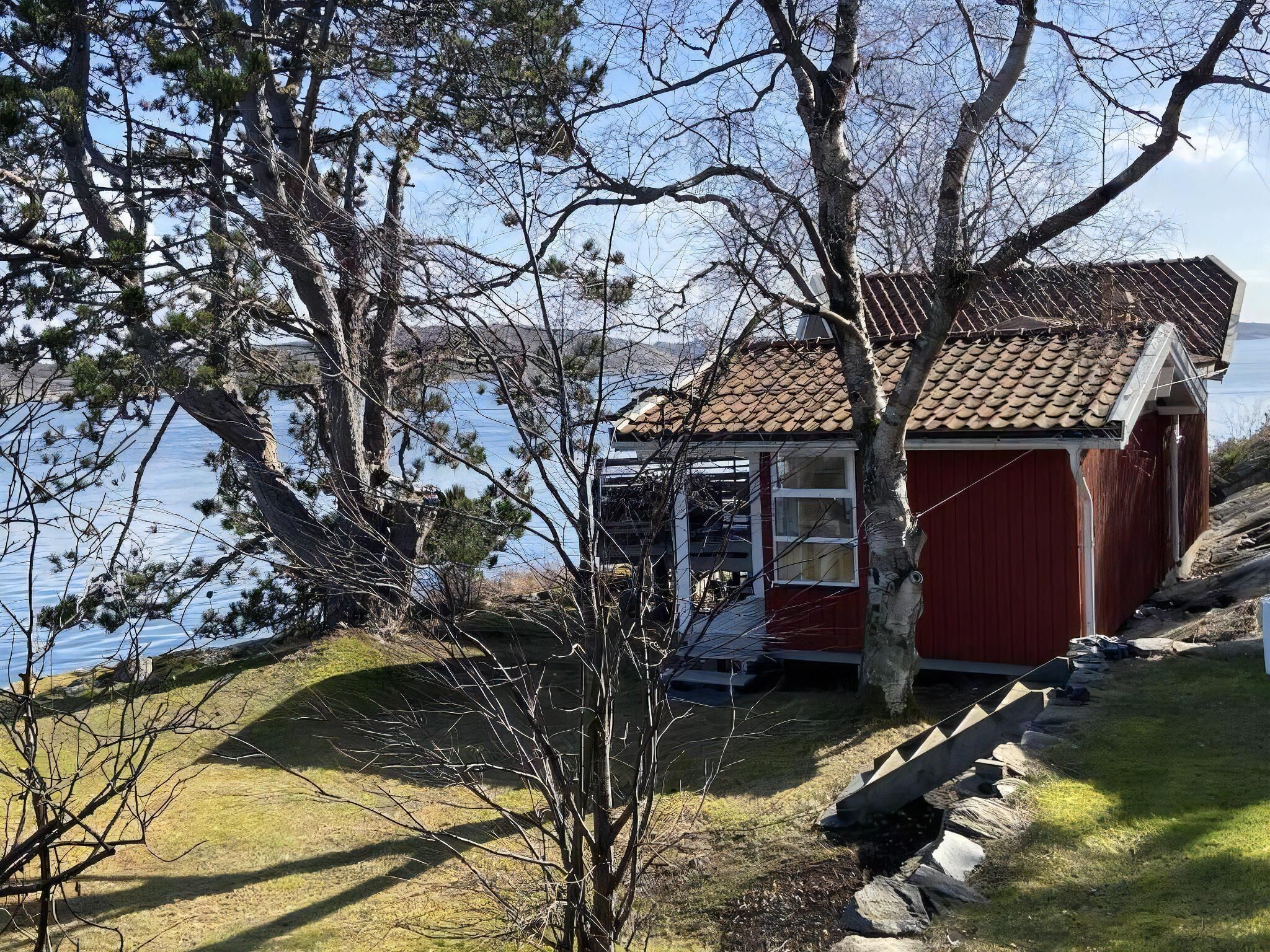 Un petit bâtiment rouge avec un toit en tuiles, situé au bord d’une étendue d’eau, entouré d’arbres et d’une zone herbeuse.