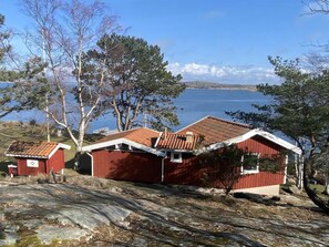 En röd trästuga med ett tak belagt med takpannor, belägen på en bergig plats med utsikt över en sjö och avlägsna kullar.