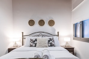 A neatly made bed with a white duvet, two pillows with floral patterns, and a rolled towel on the bed. There are two bedside tables with lamps, a metal bed frame, and three woven wall decorations.