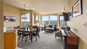 A spacious living area with a dining table, chairs, a sofa, and a coffee table. Large windows offer a view of the outdoors. - New Listing! River Views | Hot Tub | Near Mt. Bachelor (Bend)