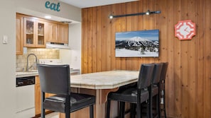 A kitchen with wooden cabinets, a dining table with chairs, a painting of a mountain, and a clock on the wall. - New! Remodeled Condo | Pool - Hot Tub | Steps to River Trail (Bend)