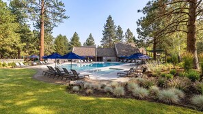 A pool area with lounge chairs and umbrellas, surrounded by trees and a building with a stone facade. - New! Remodeled Condo | Pool - Hot Tub | Steps to River Trail (Bend)