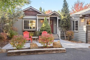 Ein zweistöckiges Haus mit Veranda, einer roten Tür und einem kleinen Garten mit einer hölzernen Bank und zwei roten Adirondack-Stühlen.