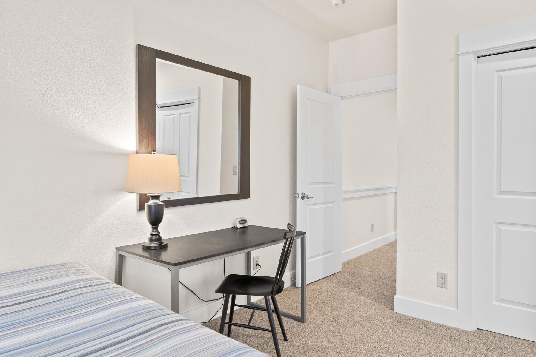 A hotel room with a bed, a desk with a chair, a lamp, and a mirror. There are two white doors and a carpeted floor.