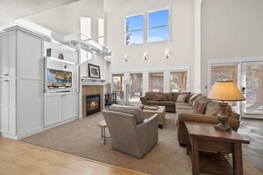 A spacious living room with a fireplace, a television, and a large window providing natural light. - New! Luxury Forest Home | Arcade Room | Pool - Hot Tub (Bend)