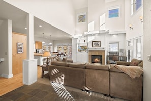 A spacious living area with a large sectional sofa, a fireplace, and a kitchen area with a dining table and chairs. - New! Luxury Forest Home | Arcade Room | Pool - Hot Tub (Bend)
