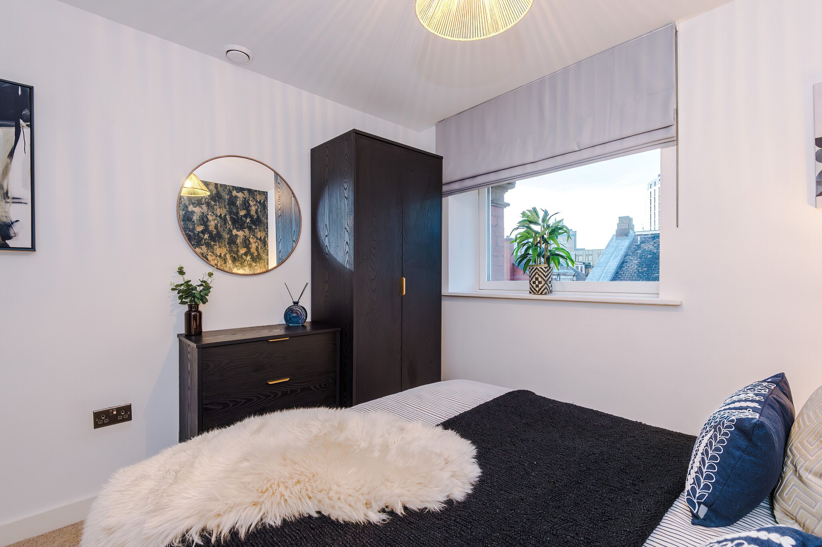 A bedroom with a large bed, a dark wardrobe, a round mirror, a window with a view of buildings, and a small plant on the dresser.