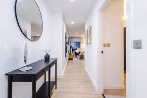 A hallway with a round mirror, a black console table, and a wooden floor leading to a living area with a sofa and a TV. - Modern 2BR Apartment Leeds Centre (Leeds)