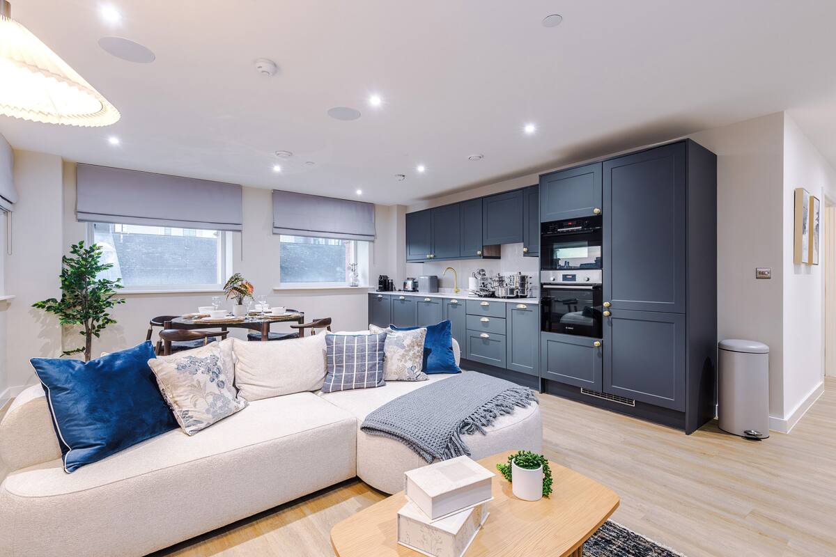 A modern kitchen with dark cabinets, a dining area with a table and chairs, a sofa with decorative pillows, and a small plant on a wooden table.