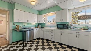 A kitchen with teal walls, white cabinets, a black and white checkered floor, a stainless steel dishwasher, and a window with a view of trees.