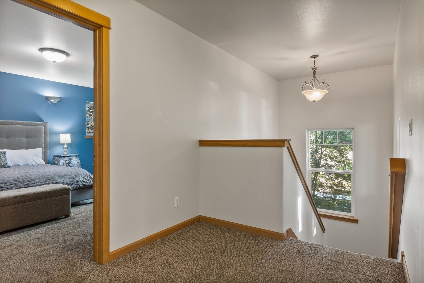 A bedroom with a bed, nightstand, and lamp; a hallway with a staircase; and a window with a view.