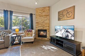 Ein Wohnzimmer mit Kamin aus Stein, einer Couch, einem Sessel, einem Fernseher und einem Fenster mit Vorhängen.