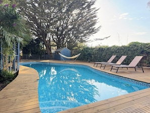 A swimming pool with a wooden deck, lounge chairs, a hammock, and a large tree. - Palm House | Pool, Hot Tub | South Downs (Worthing)