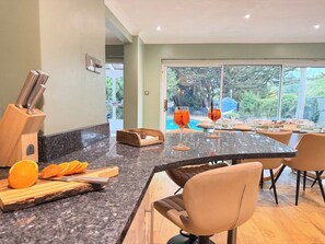 A modern kitchen with a granite countertop, a wooden fruit bowl, and a dining area with a glass table and chairs. - Palm House | Pool, Hot Tub | South Downs (Worthing)