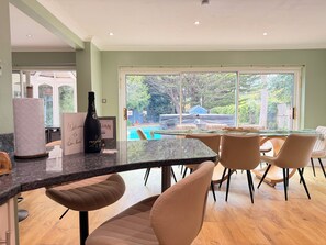 A modern kitchen with a large island, a dining area with chairs, and a view of a swimming pool. - Palm House | Pool, Hot Tub | South Downs (Worthing)