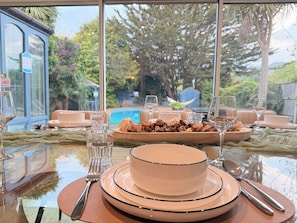 A dining table set with plates, glasses, and cutlery, with a pool and trees visible through the window. - Palm House | Pool, Hot Tub | South Downs (Worthing)
