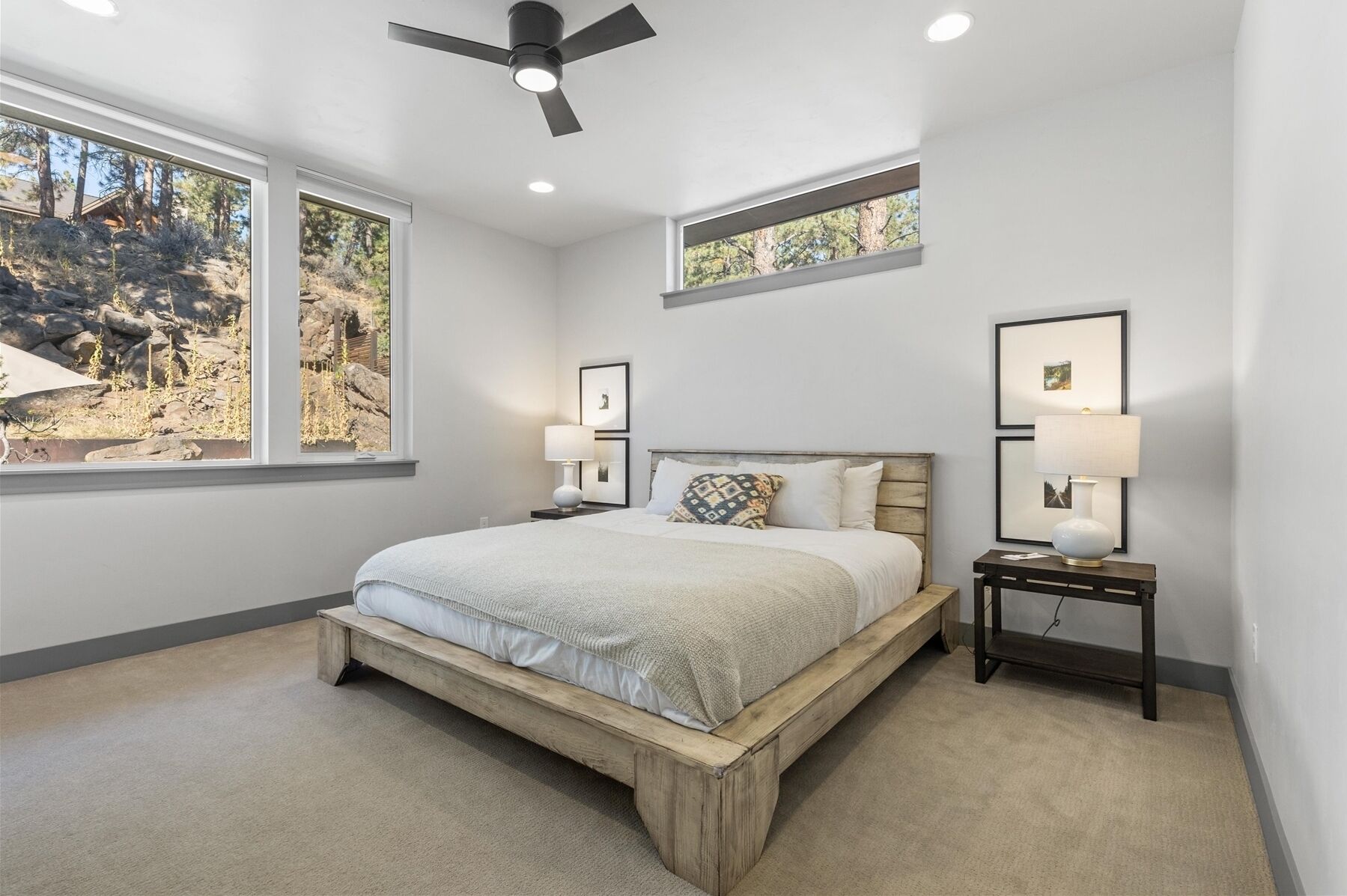 A bedroom with a large bed, two bedside tables, a ceiling fan, and two windows with a view of trees.