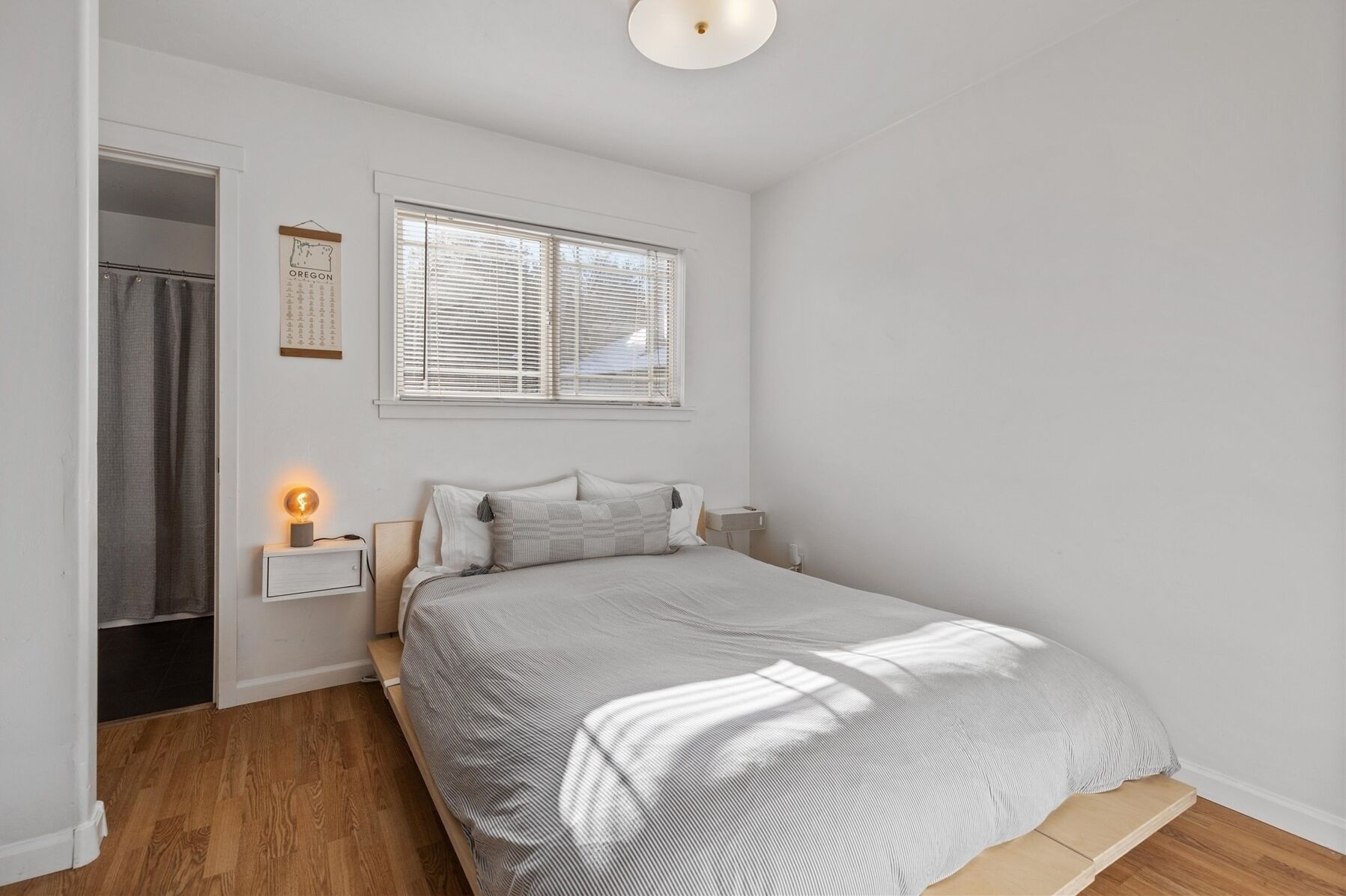 Een slaapkamer met een bed, een nachtkastje, een raam met jaloezieën, een deur en een wand met een lijst erin.