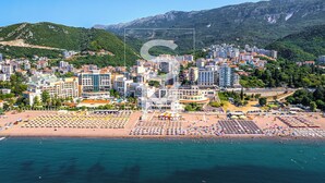Un paisaje urbano costero con una zona de playa que cuenta con numerosas tumbonas y sombrillas, rodeada de edificios residenciales y montañas al fondo.