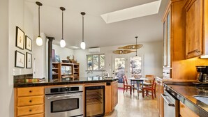 Cocina moderna con una isla central, hornos empotrados y alacenas de madera. Hay luces colgantes, una zona de comedor con sillas y vistas a través de las ventanas.