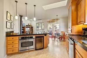 Eine moderne Küche mit einer zentralen Kochinsel, eingebauten Backöfen und Holzschränken. Es gibt Pendelleuchten, einen Essbereich mit Stühlen und einen Ausblick durch die Fenster.