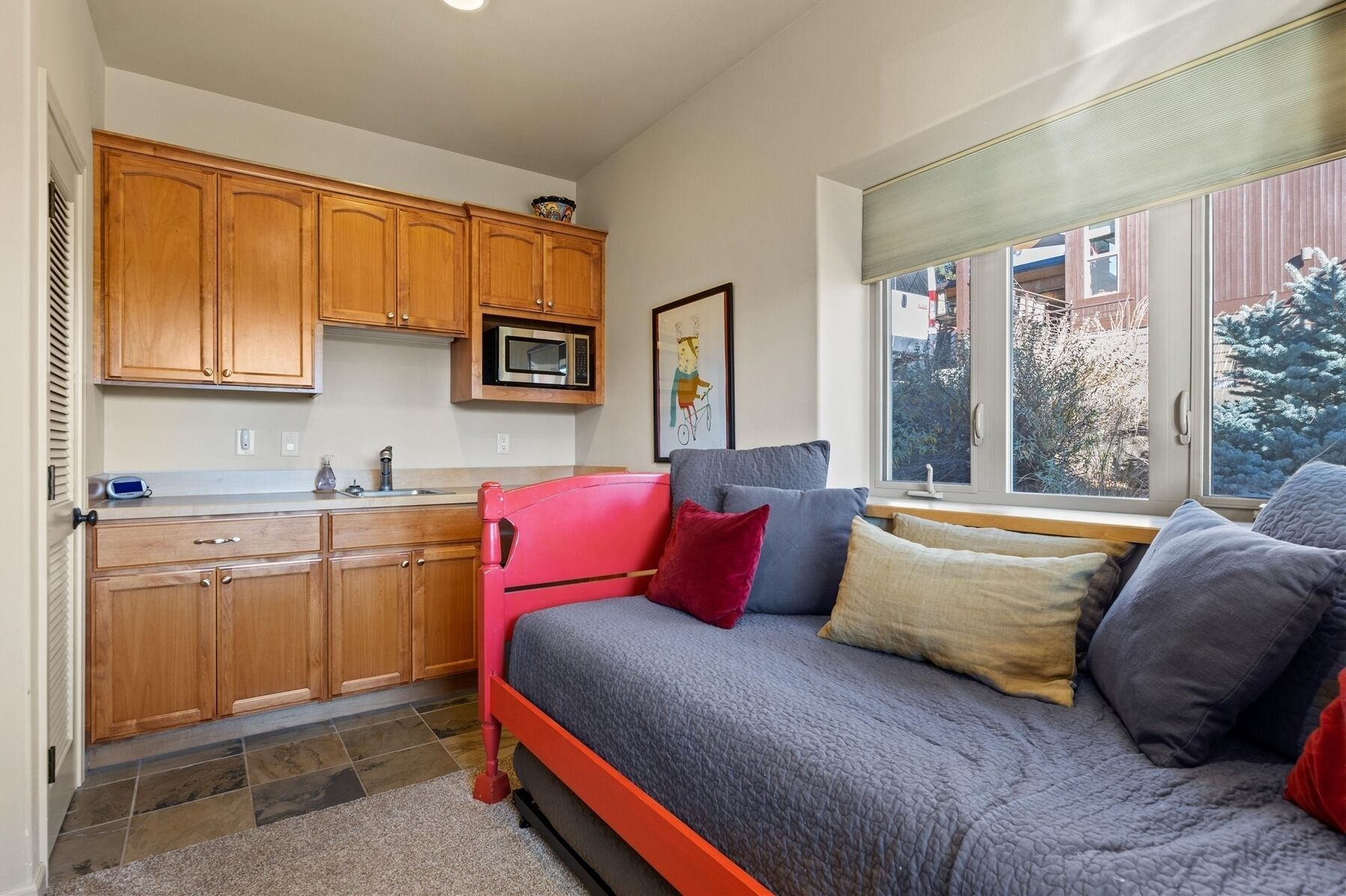 Un espacio habitable compacto con kitchenette, una cama con muchas almohadas y un ventanal con vistas a árboles.