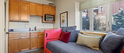 Un espacio habitable compacto con kitchenette, una cama con muchas almohadas y un ventanal con vistas a árboles.