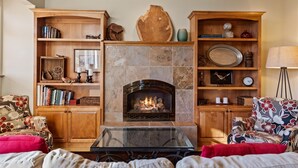 Una sala de estar con chimenea, estanterías de madera, una mesa de centro de vidrio y muebles con estampado floral.