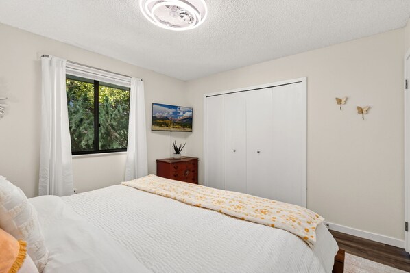 Un dormitorio con cama, ventana con cortinas, cómoda, puerta y un cuadro colgado en la pared.