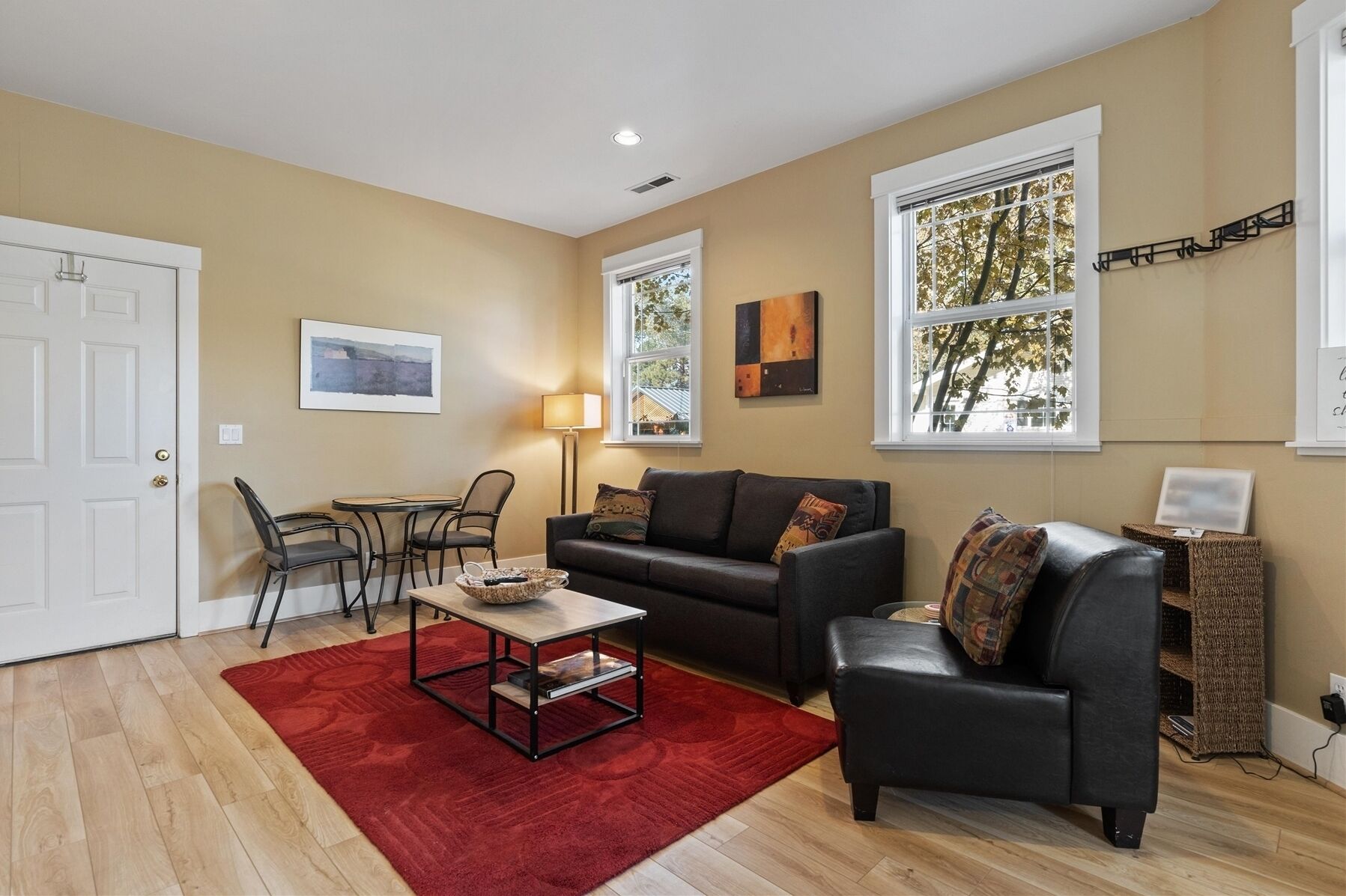 A living room with a sofa, a coffee table, a red rug, and a lamp. There are two windows with curtains, a door, and a wall with artwork.