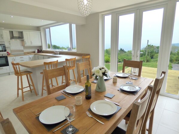 A modern kitchen with white cabinets, a wooden dining table set for four, and large windows offering a view of the outdoors. - Modern Seaside house, walking distance to beautiful beach and coastal path (freshwater east)