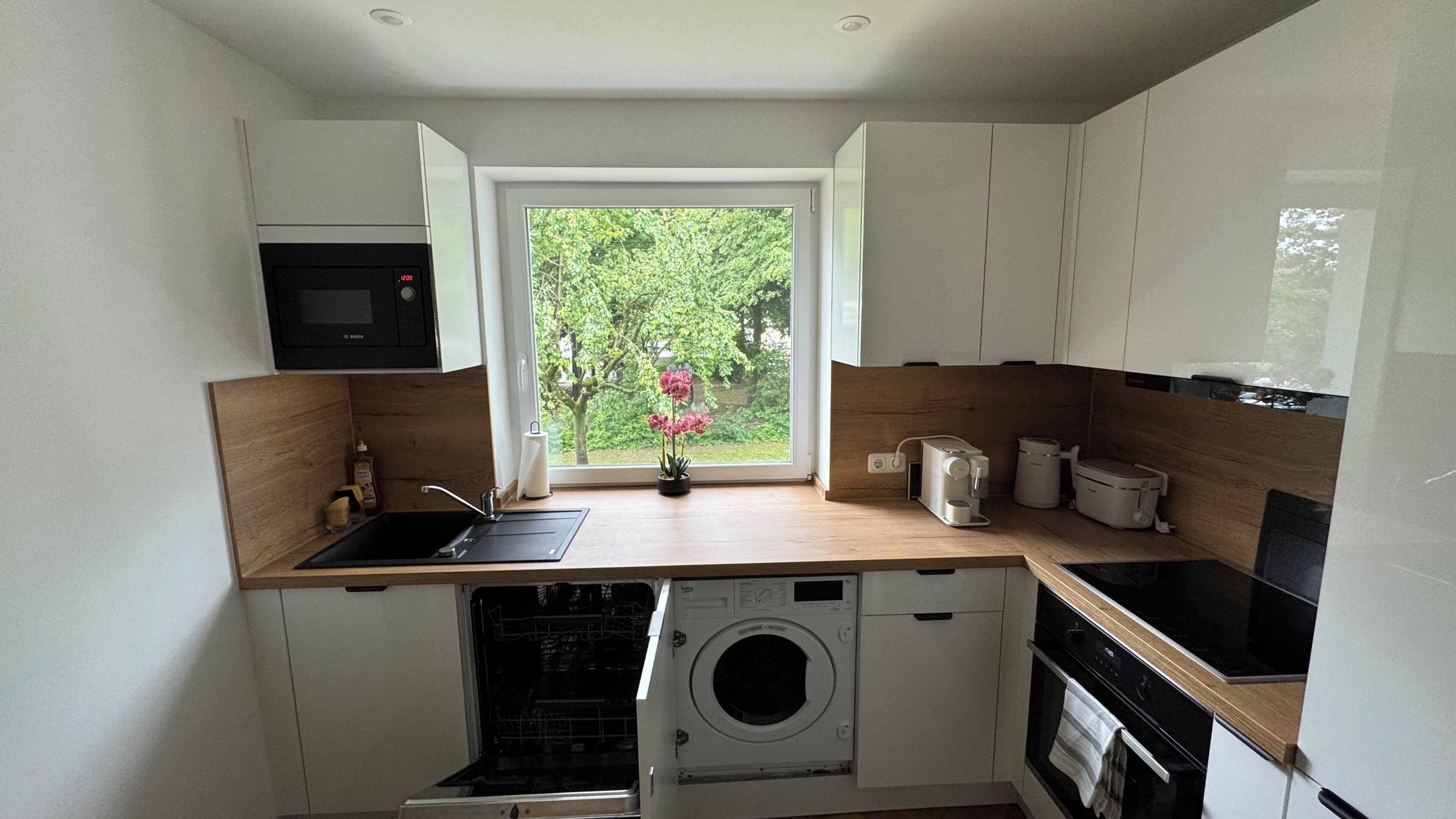 A compact kitchen with a washing machine, a microwave, and a window with a view of greenery.