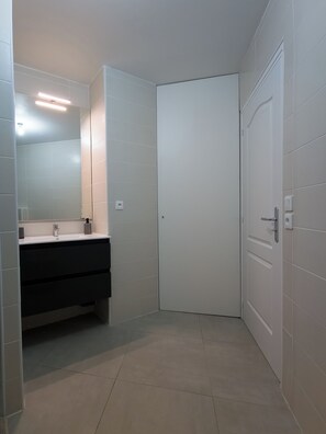 Ein Badezimmer mit weiß gefliesten Wänden, einem dunklen Waschtisch, einem Spiegel, einer weißen Tür und einer Leuchte.
