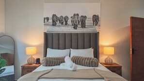 A bedroom with a large bed, two bedside tables, a wall-mounted lamp, and a poster of elephants.