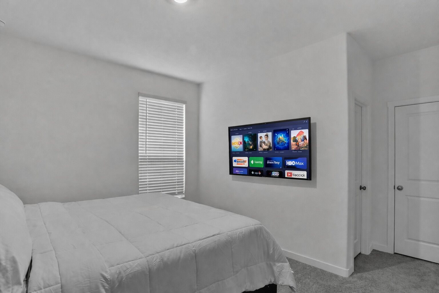 Une chambre à coucher avec un lit, un téléviseur fixé au mur, une fenêtre avec des stores et une porte.