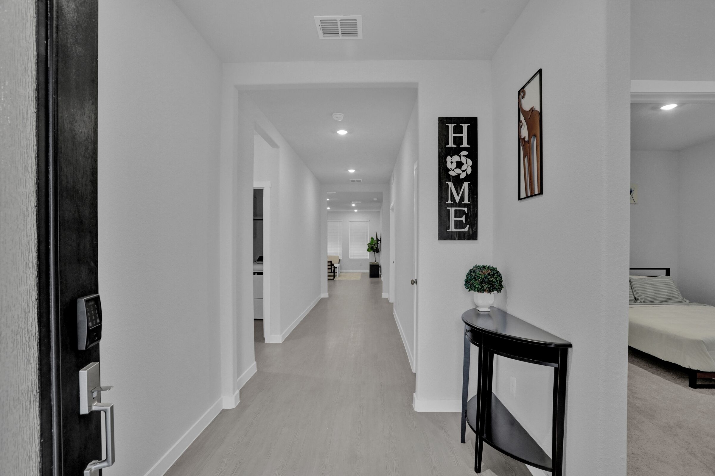 Un couloir avec une pancarte indiquant « MAISON », un bureau avec une plante et un lit à l’arrière-plan.