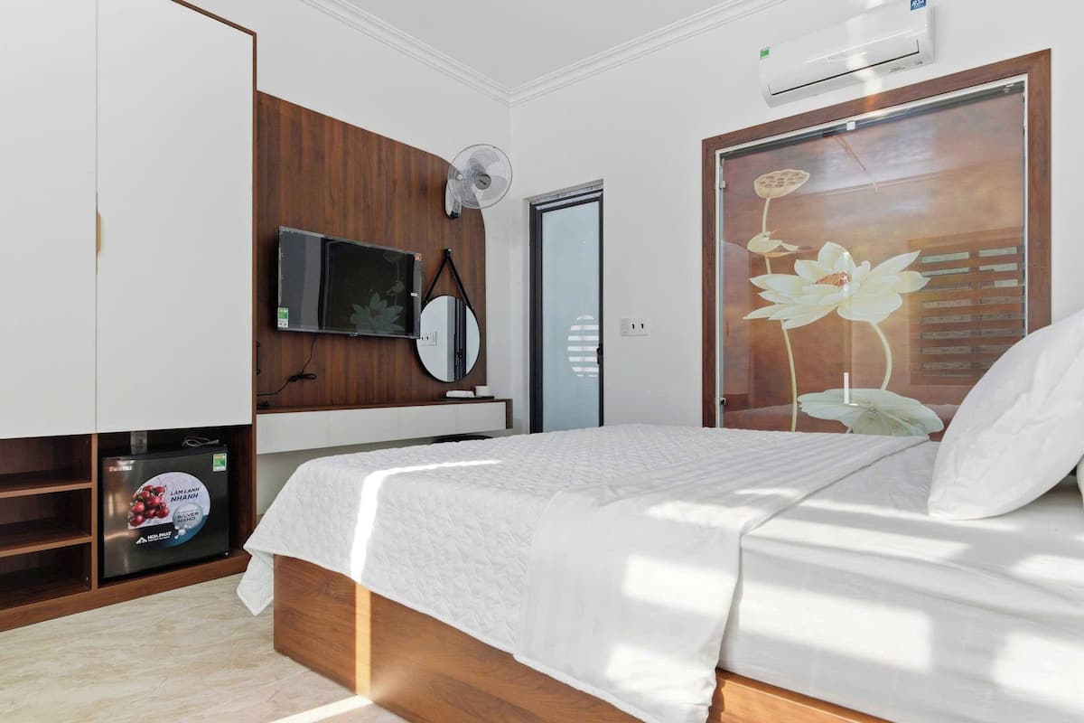Une chambre à coucher avec un lit, un téléviseur fixé au mur, un ventilateur et un réfrigérateur.